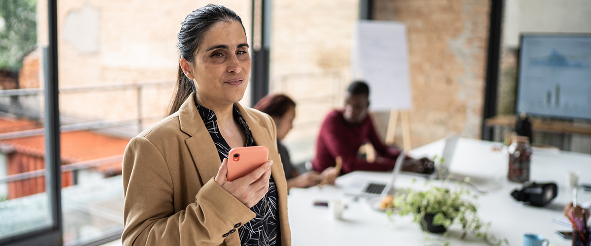 A middle aged women with long dark brown in a ponytail, wearing a business style suit standing in front of a table at a presentation using her cell phone for dictation of a website.
